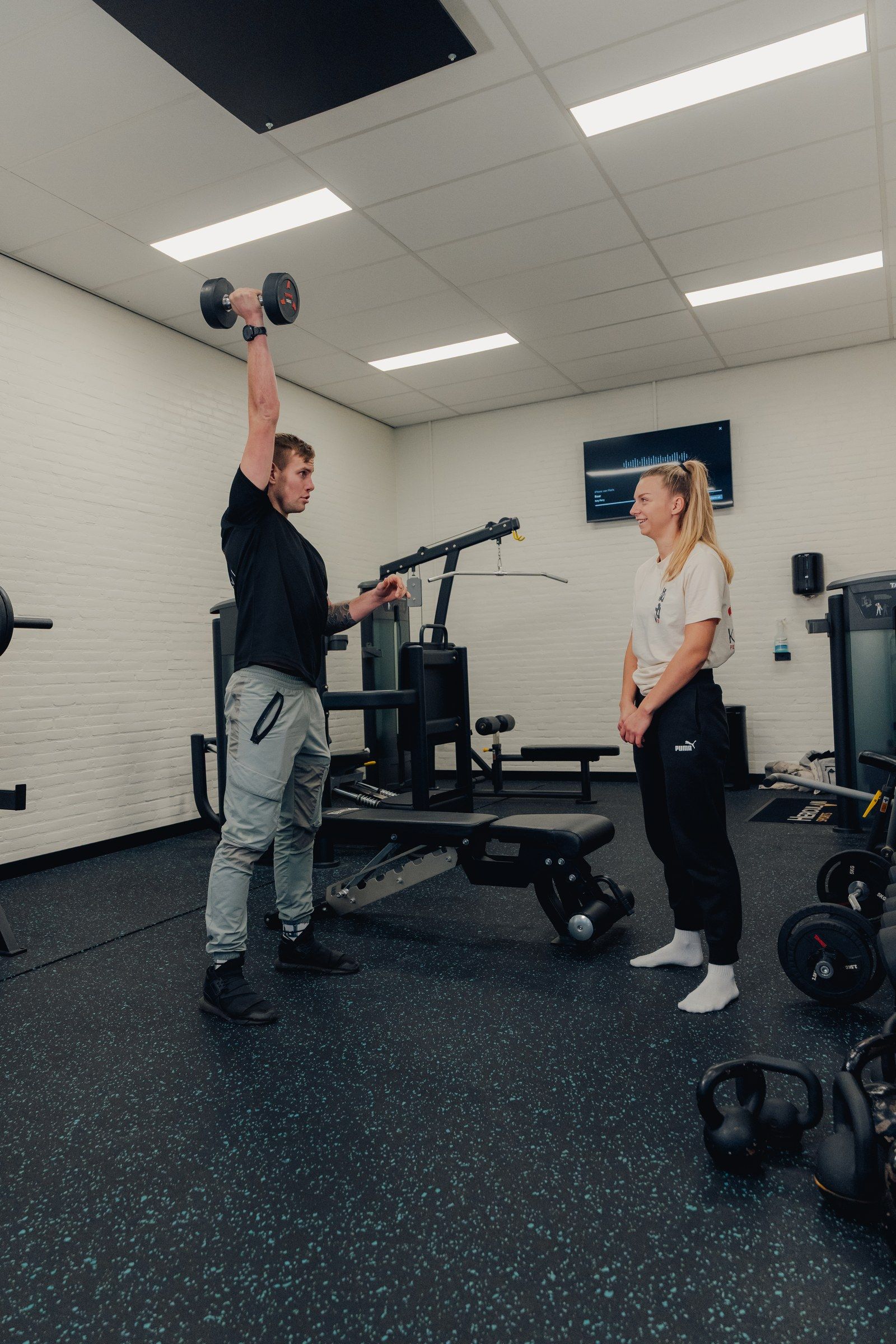Floris drukt een dumbbell boven zijn hoofd en corrigeert een lachende klant in de gym.