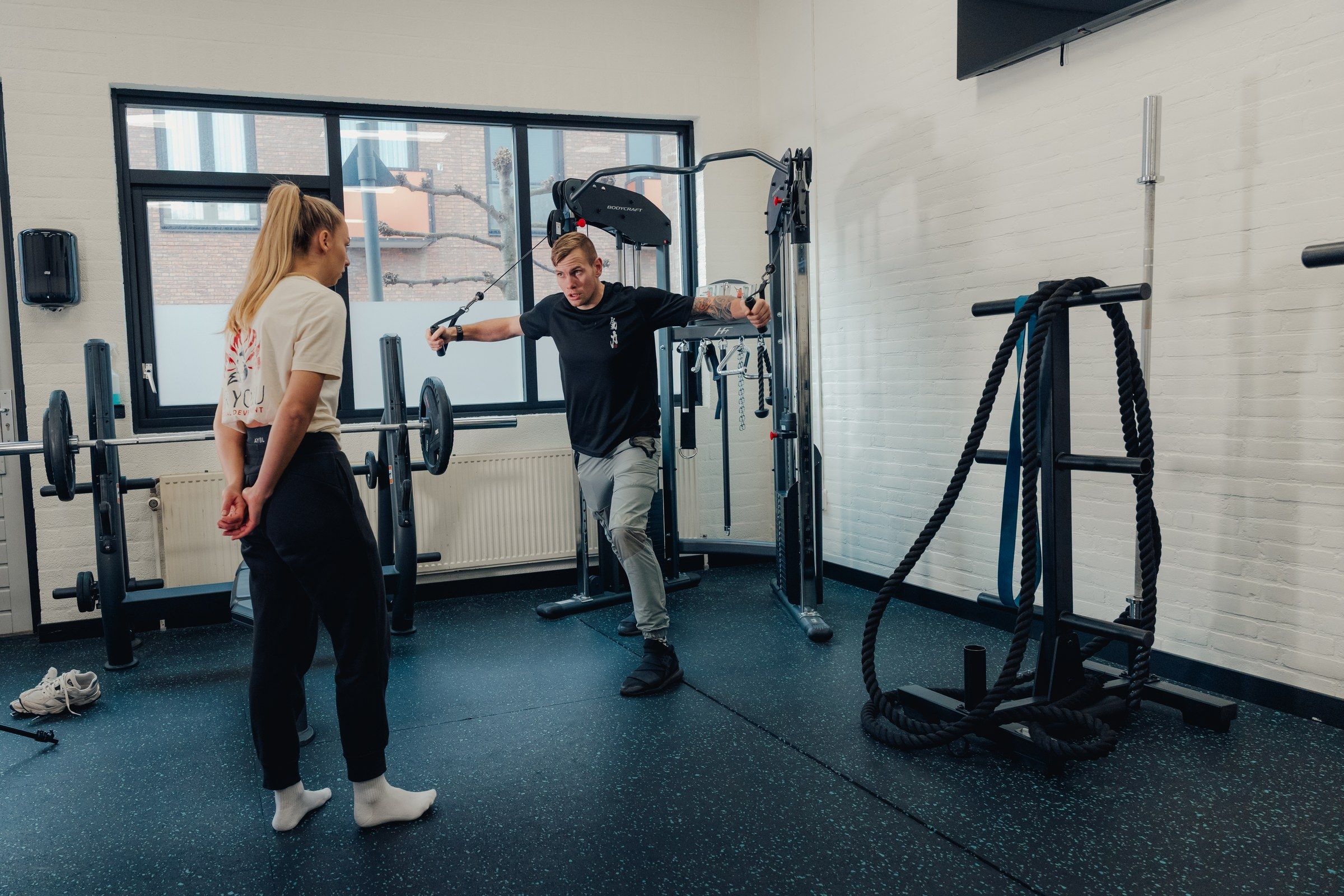 Floris doet in een lichte gym met een groot raam een kabeltrek-oefening voor; een klant kijkt toe.