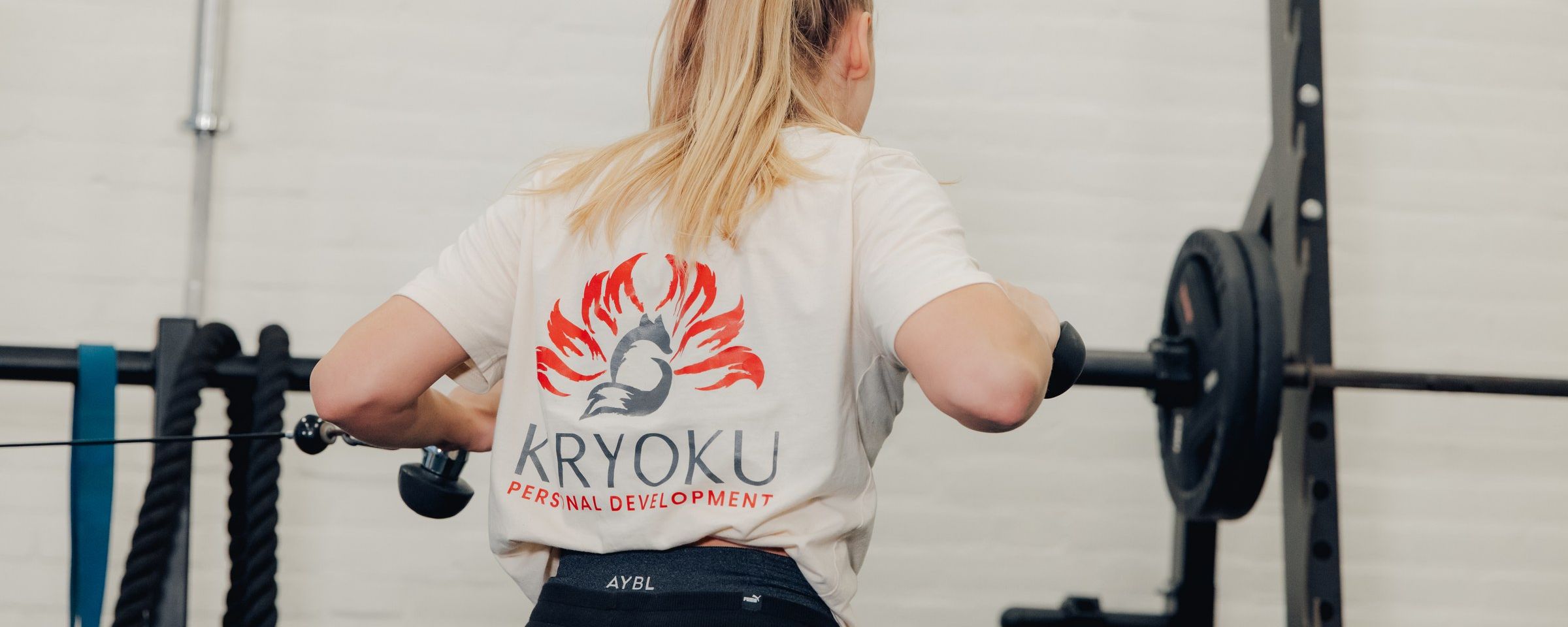 Een klant in een crème Kiryoku-shirt met het vos-logo op de rug, bij een rack in de gym.